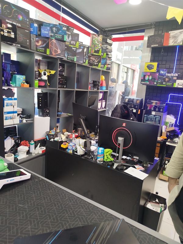 Balcão da loja com monitores e, ao fundo, prateleiras com caixas de placas de vídeo e fontes