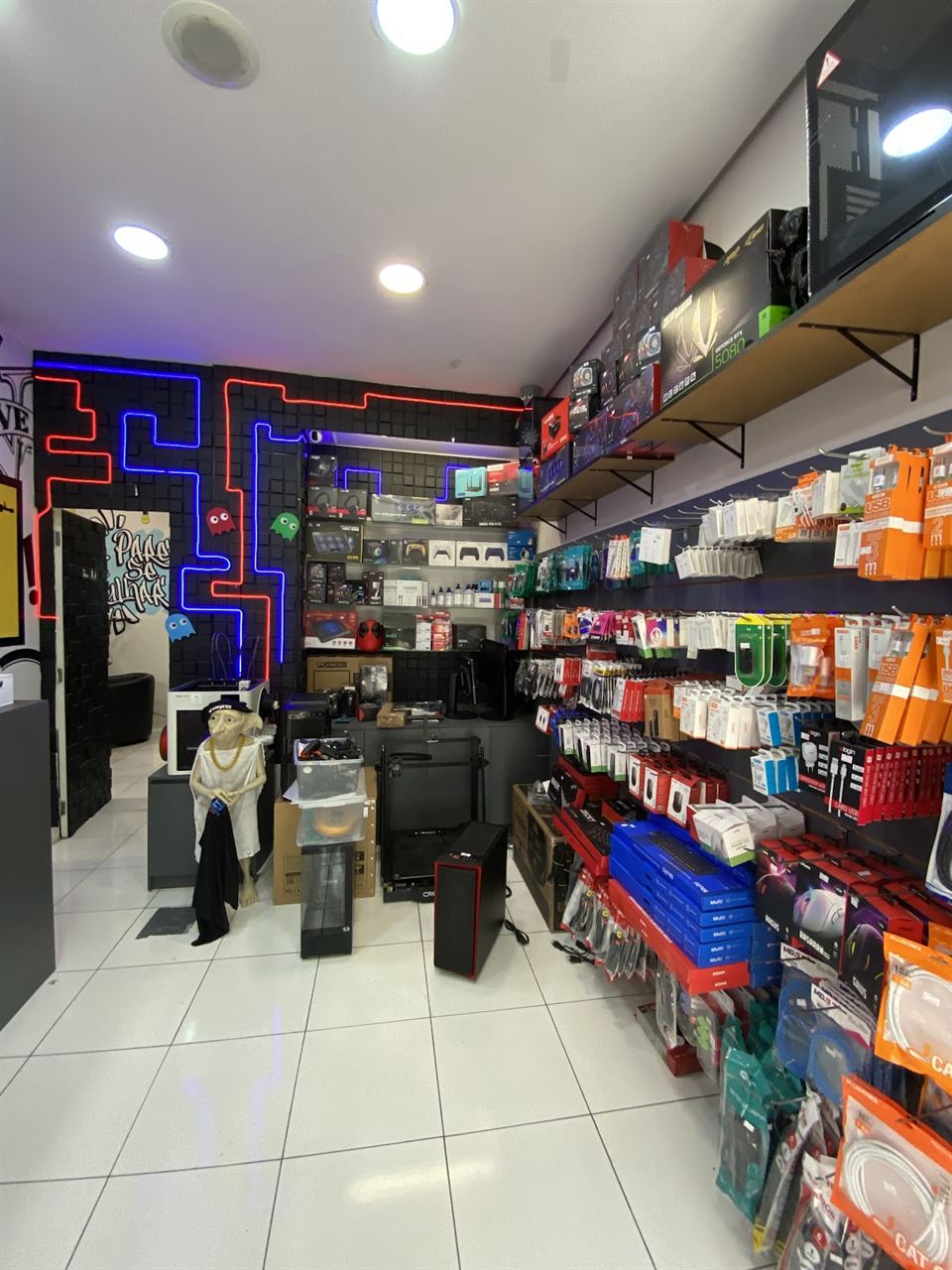 Interior da Benincasa Info no Centro Comercial Alphaville, com o labirinto de neon azul e vermelho na parede do fundo e a prateleira de consoles e acessórios