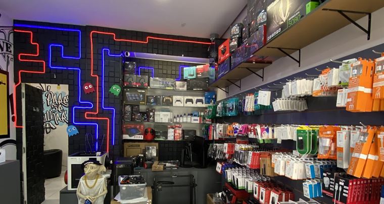 Interior da Benincasa Info, com o labirinto de neon azul e vermelho na parede e prateleiras de consoles e acessórios